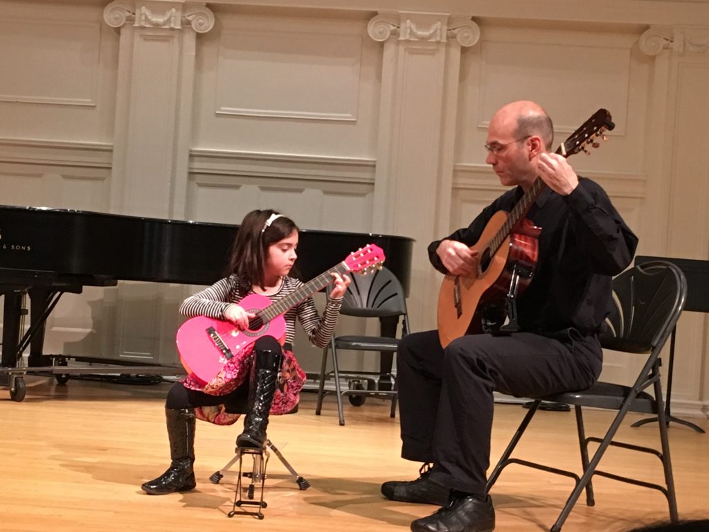 Teacher sits next to a Young girl with a pink classical guitar with a piano in the background.
